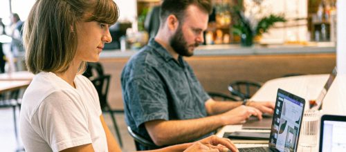 Two young professionals working on laptops in a modern cafe setting.